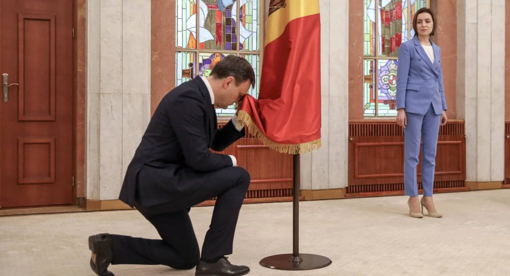 New Moldovan Prime Minister Dorin Recean kneels down in front of a state flag as President Maia Sandu stands nearby during an inauguration ceremony in Chisinau, Moldova, February 16, 2023. Photo credit: Reuters